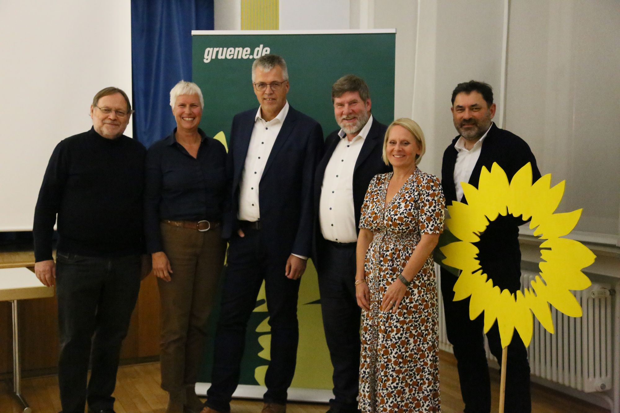 Zwei Frauen und vier Männer stehen vor einer Tafel und lachen in die Kamera. Seitlich ist eine stilisierte Sonnenblume. 