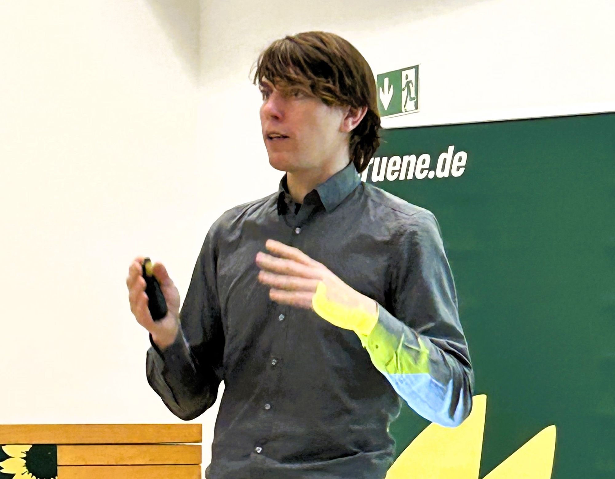 Ein junger Mann steht vor einer Tafel und spricht unter zu Hilfenahme von beiden Händen.