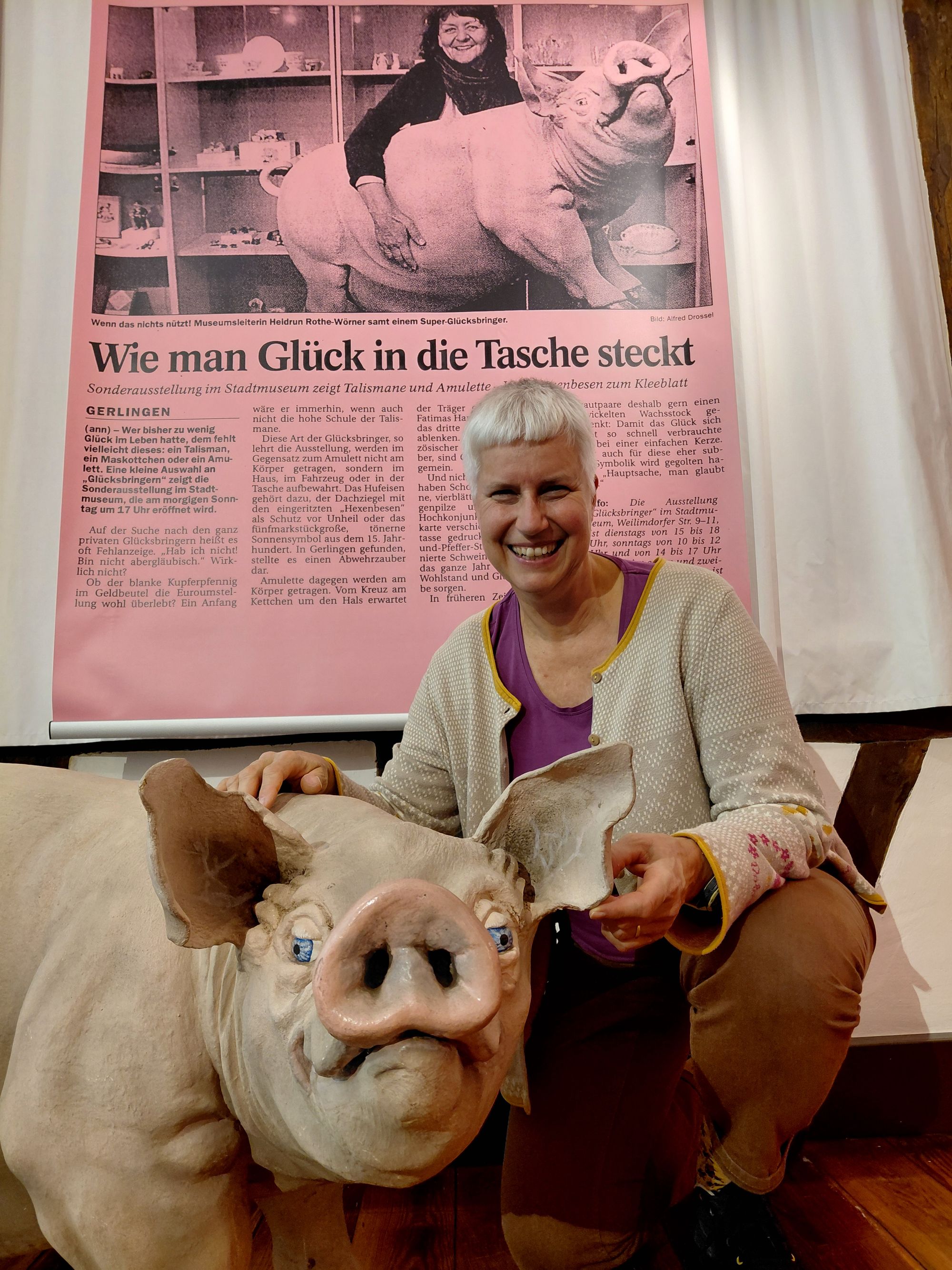 Im Vordergrund steht ein großes Glücksschwein. Daneben kniet die Autorin des Fotos. Im Hintergrund ist ein Zeitungsausschnitt zu sehen, auf dem die frühere Museumsleiterin Frau Rothe das gleiche Glücksschwein im Arm hält.