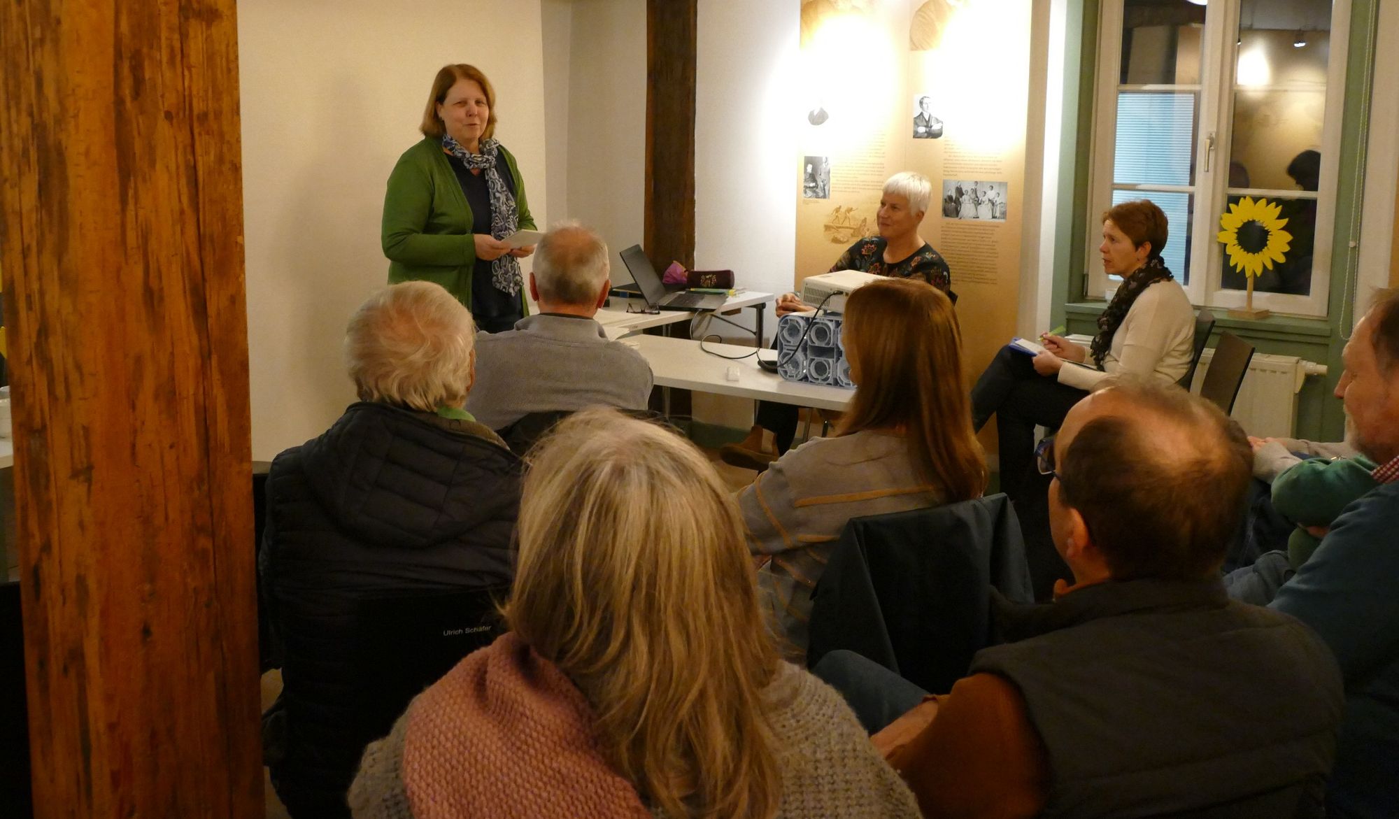 Eine Frau in grüner Jacke steht vor mehreren sitzenden Zuhörern und Zuhörerinnen, die nur von hinten zu sehen sind. 