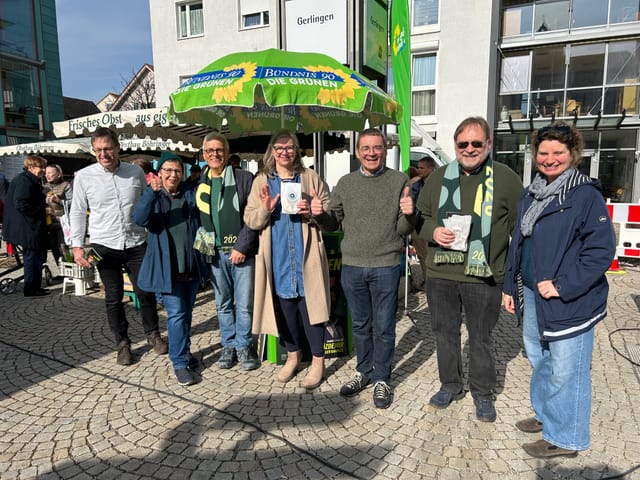 Sieben Personen stehen auf einem Platz unter einem grünen Sonnenschirm 